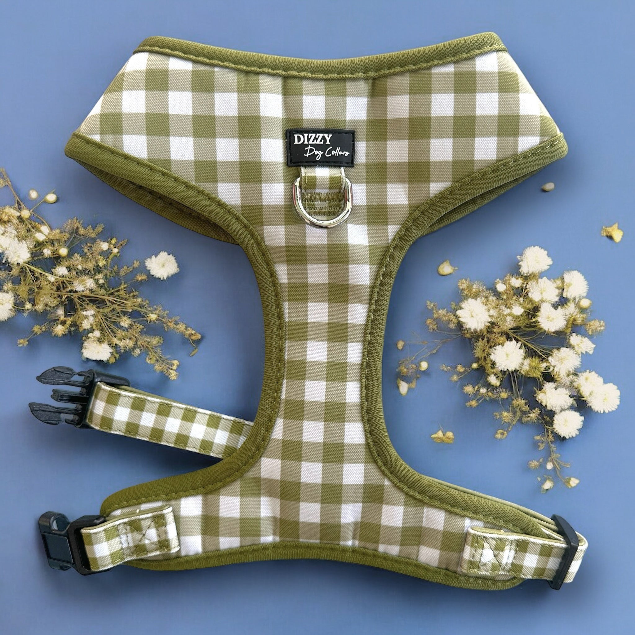The image showcases a green and white checkered dog harness on a light blue background, accented by white and gold flowers. The harness is labeled