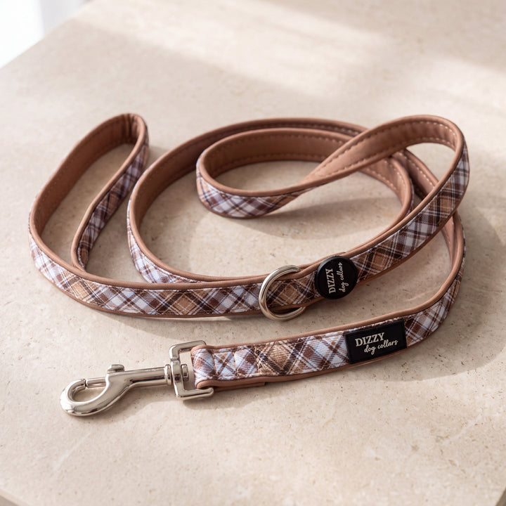 A dog leash with a brown and beige plaid pattern and tan leather trim, coiled on a light beige stone surface. It features a silver metal clasp, a black circular brand tag, and a black fabric brand label reading 'DIZZY by dizzymani'.